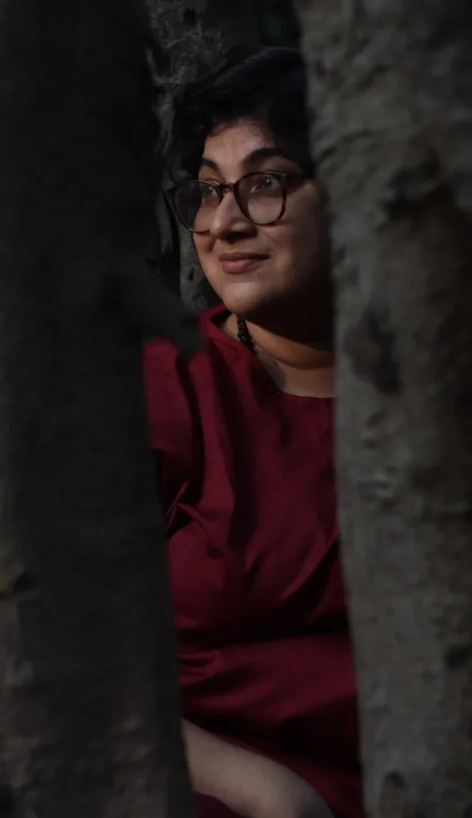A person in red sits partially hidden among the thick trunks of clustered trees, their face turned toward the light breaking through the canopy.