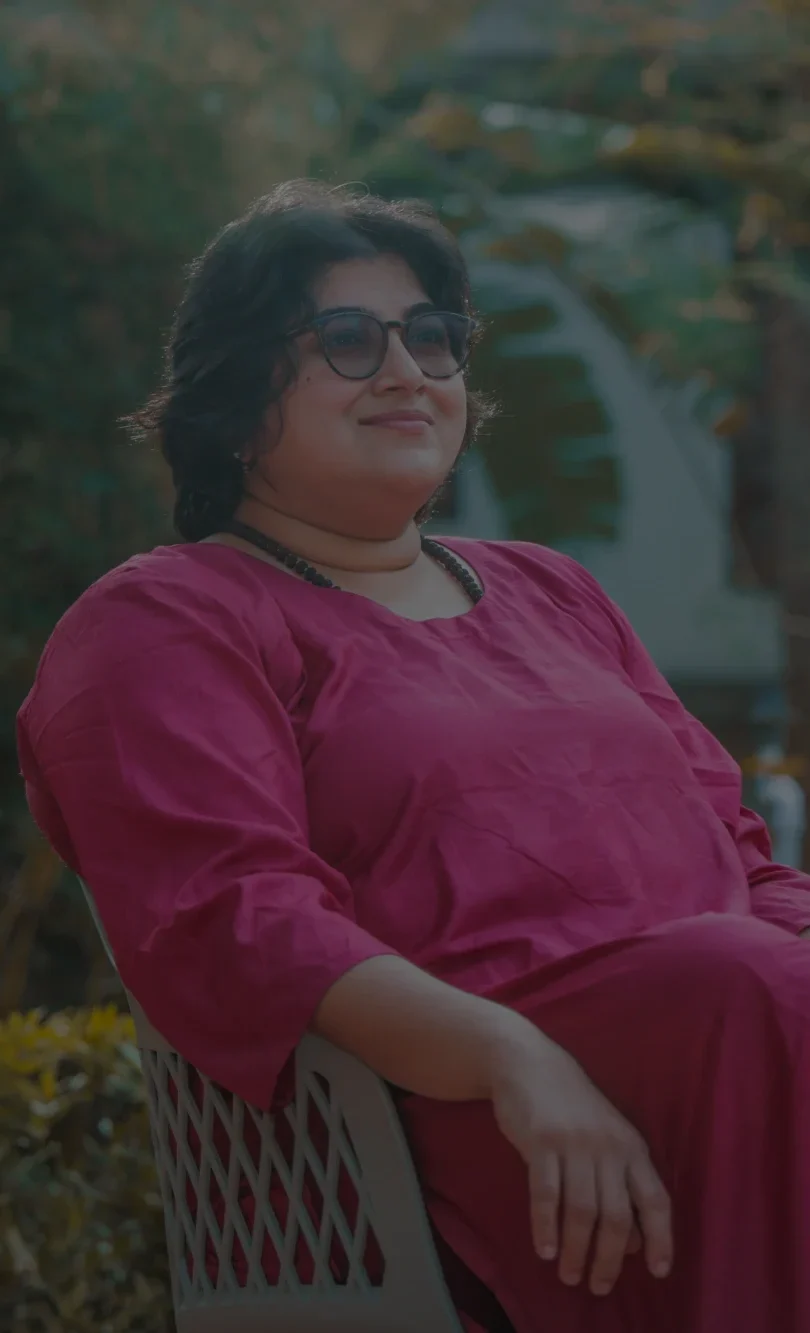 A person sits outdoors in a chair, wearing a long red co-ord and glasses, with sunlight filtering through trees behind them. They rest one arm on their lap and look slightly upward.