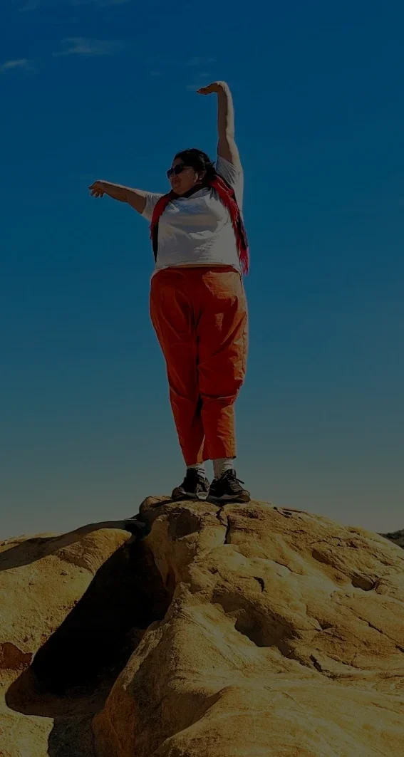 A person stands on top of a sunlit rock formation with arms lifted in an expressive pose, framed by a bright blue sky and rolling desert hills in the distance.