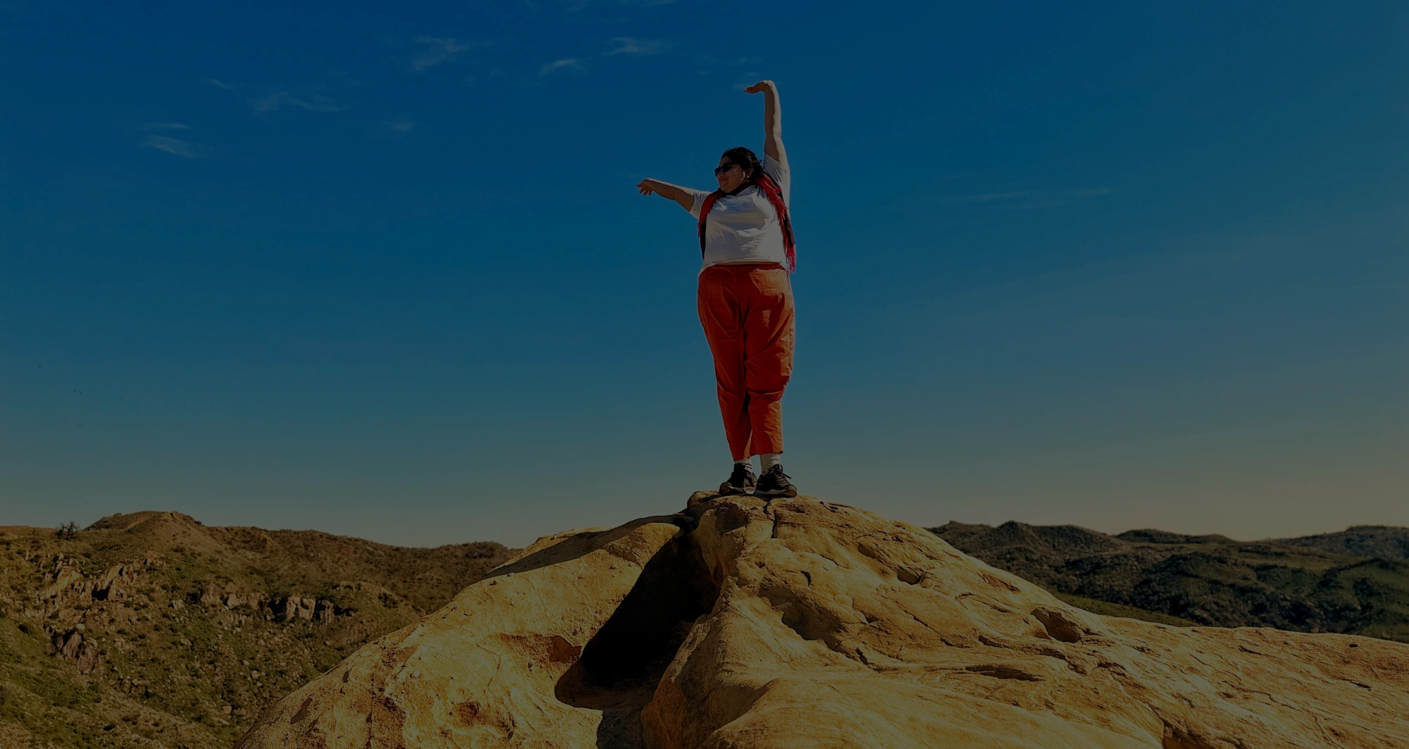 A person stands on top of a sunlit rock formation with arms lifted in an expressive pose, framed by a bright blue sky and rolling desert hills in the distance.