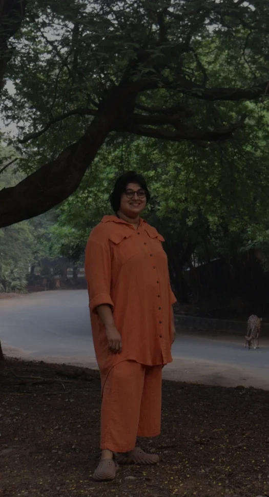 A person in an orange outfit stands under a large spreading tree beside a quiet road, while several spotted deer graze nearby in the soft morning light.