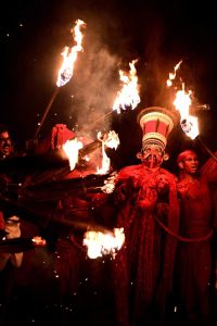 A Kathakali Ninam performer covered in vivid red stands surrounded by torch flames, their tall headgear glowing as firelight and sparks fill the dark night.