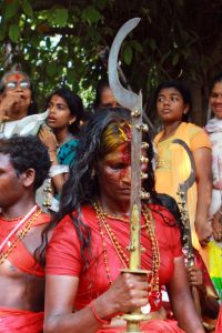 A Komaram woman from Kodungallur Bharani stands holding a sword, blood running down her forehead as onlookers watch.