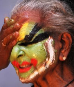 A close-up of a Kathakali performer having vivid green, red, and black makeup gently wiped from their face.