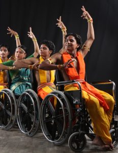 A group of dancers in wheelchairs performs Bharatanatyam hand gestures in coordinated poses, dressed in bright traditional costumes.