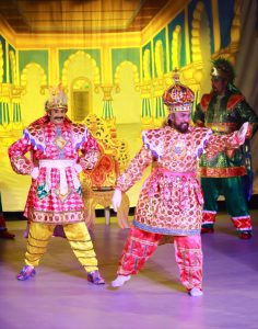 Two performers in bright, ornate Chavittu Nadakam costumes stand in bold, theatrical poses at the center. Their outfits glow with deep magenta, gold accents, and vivid blue details, framed by elaborate shoulder pieces and layered fabrics that catch the light. Other performers appear in the background.