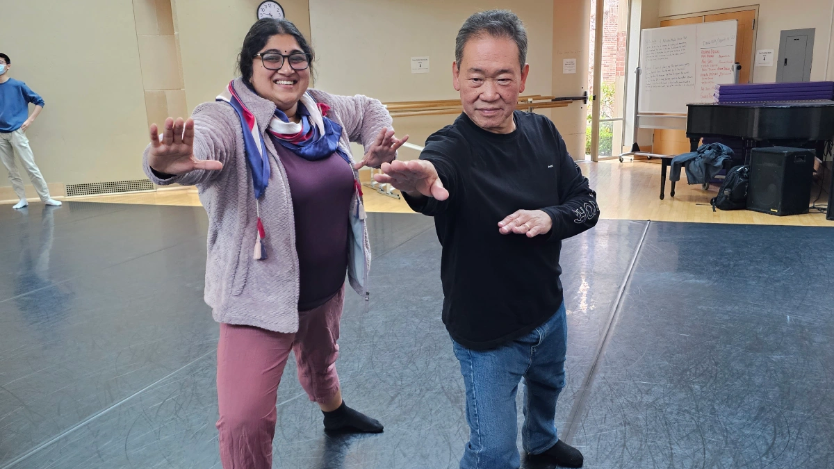 Two people stand in a dance studio striking playful martial-arts–inspired poses, both smiling as they extend their arms forward.