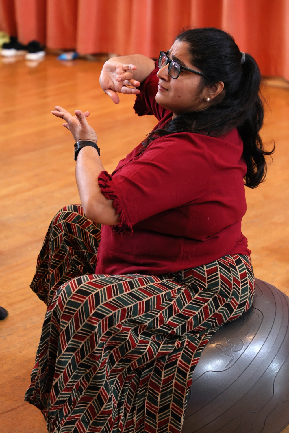 A seated dancer balances on an exercise ball, shaping a gesture with both hands, their gaze lifted as if listening through motion.