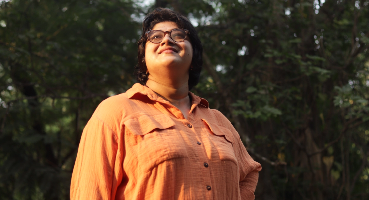 A person in an orange outfit stands outdoors, looking upward sunlight catching the edges of their face as trees blur behind them.