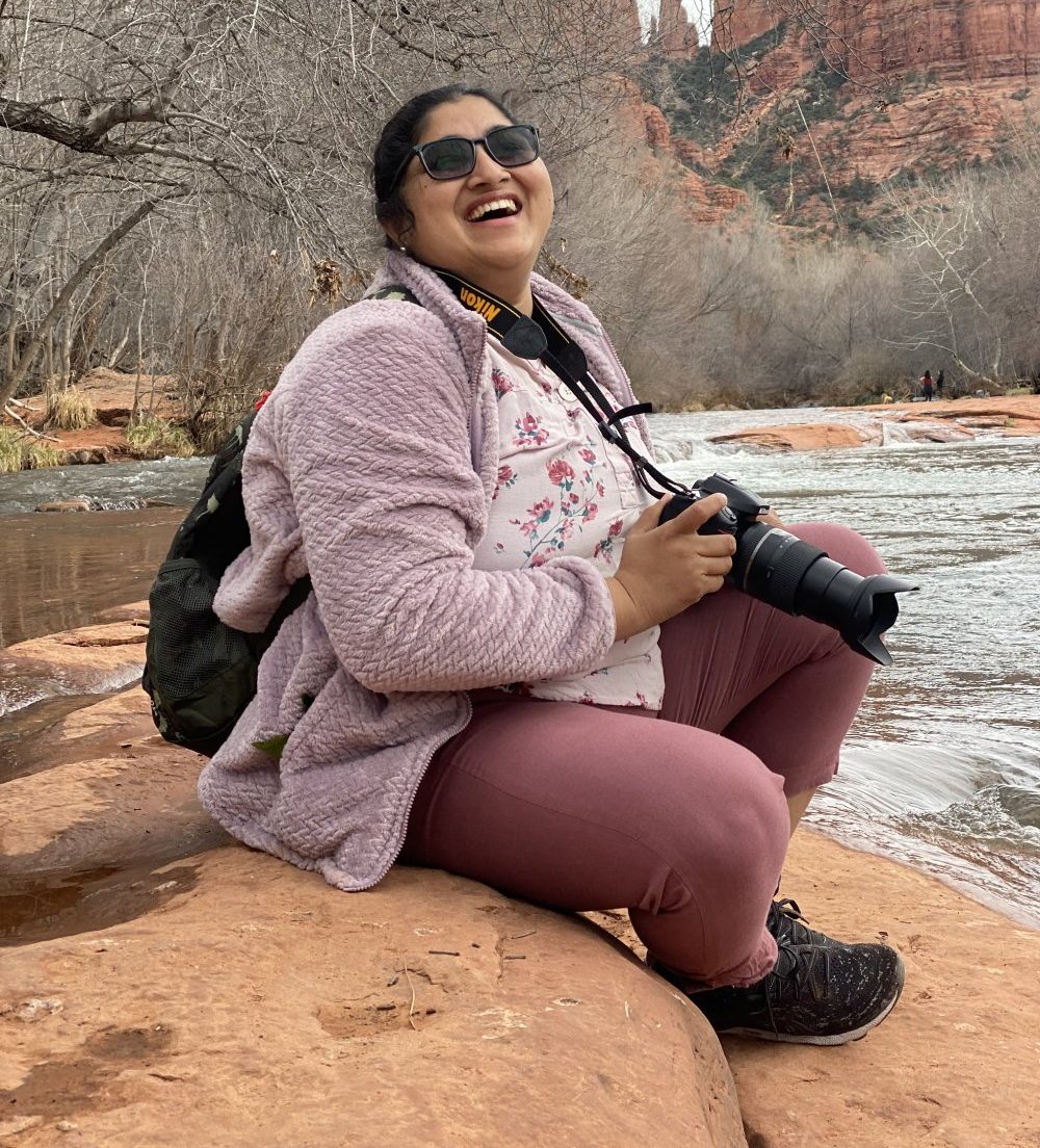 A person sitting on red rock beside a river laughs while holding a camera, with leafless trees and tall canyon walls rising in the background.