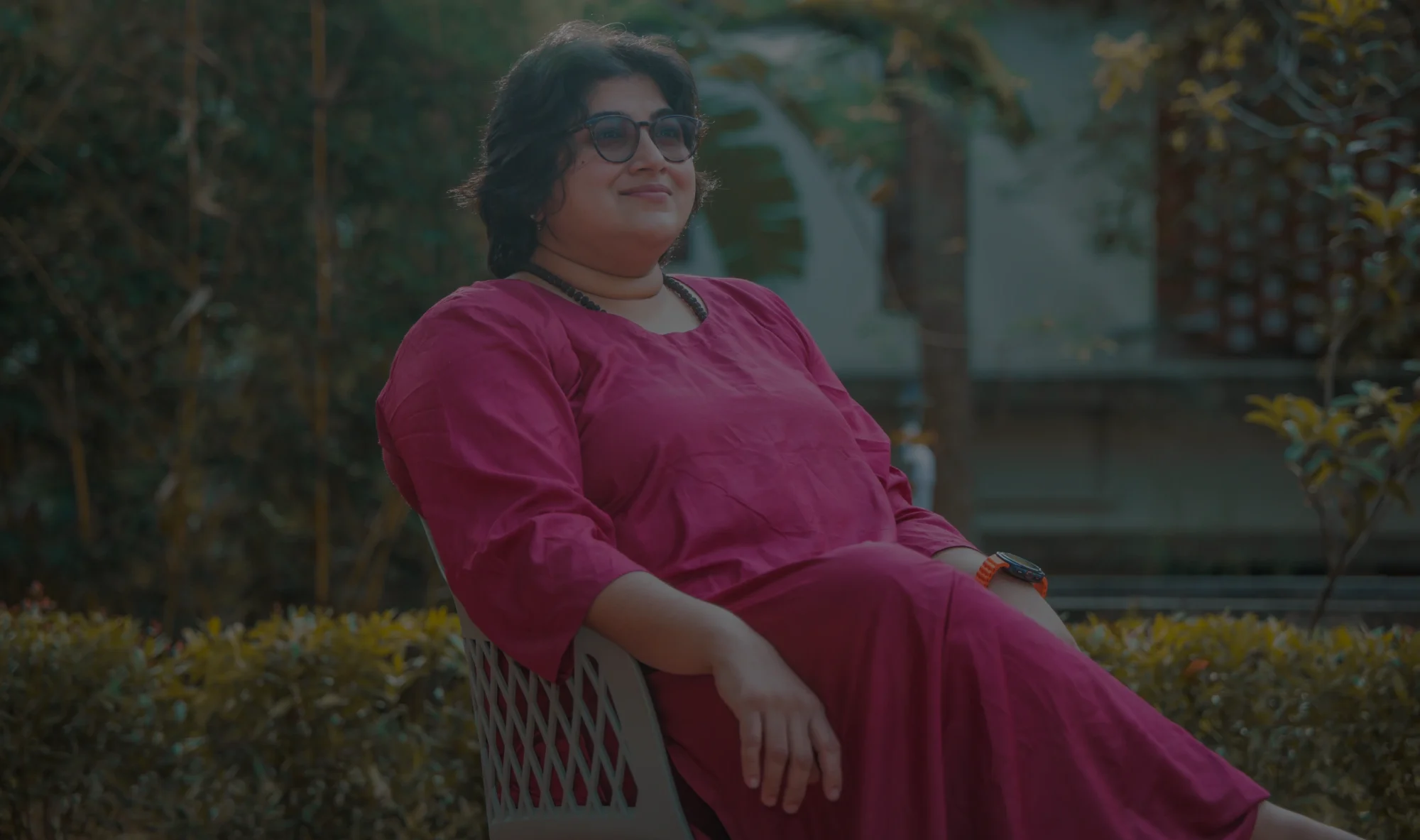 A person sits outdoors in a chair, wearing a long red co-ord and glasses, with sunlight filtering through trees behind them. They rest one arm on their lap and look slightly upward.