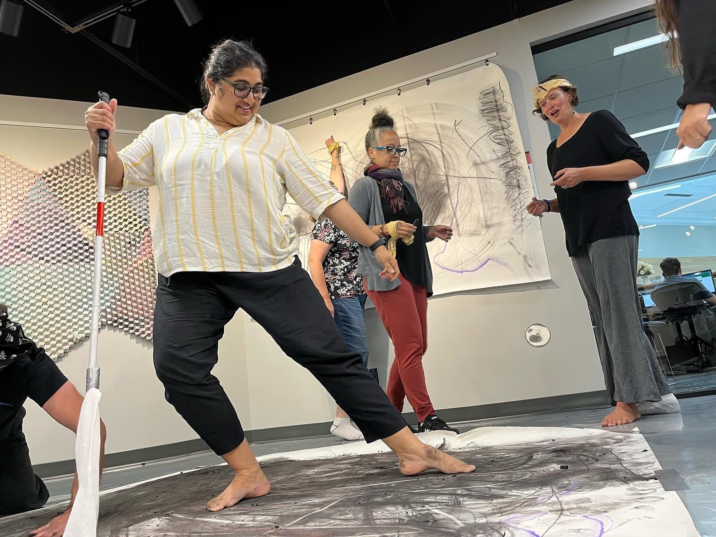 A person stands barefoot on a large sheet of charcoal-covered paper, using a white cane to guide their movement while others gather around watching and smiling.