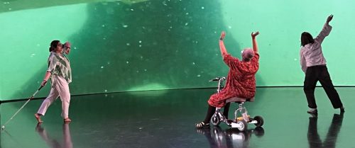 Dancers are reflected on a shiny stage floor, performing in front of a large green-tinted projection showing abstract underwater shapes. One dancer uses a cane, and another is seated on a rolling stool.