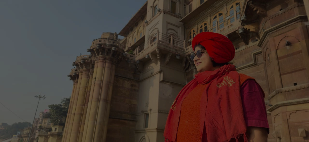 A person wearing a bright red turban and matching scarf stands near an ornate historic building with tall pillars and carved details, looking into the distance under a pale blue sky.