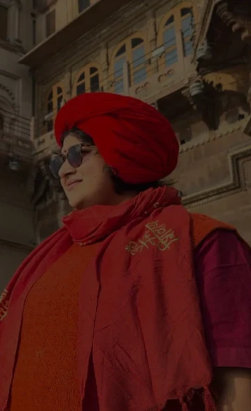 A person wearing a bright red turban and matching scarf stands near an ornate historic building with tall pillars and carved details, looking into the distance under a pale blue sky.