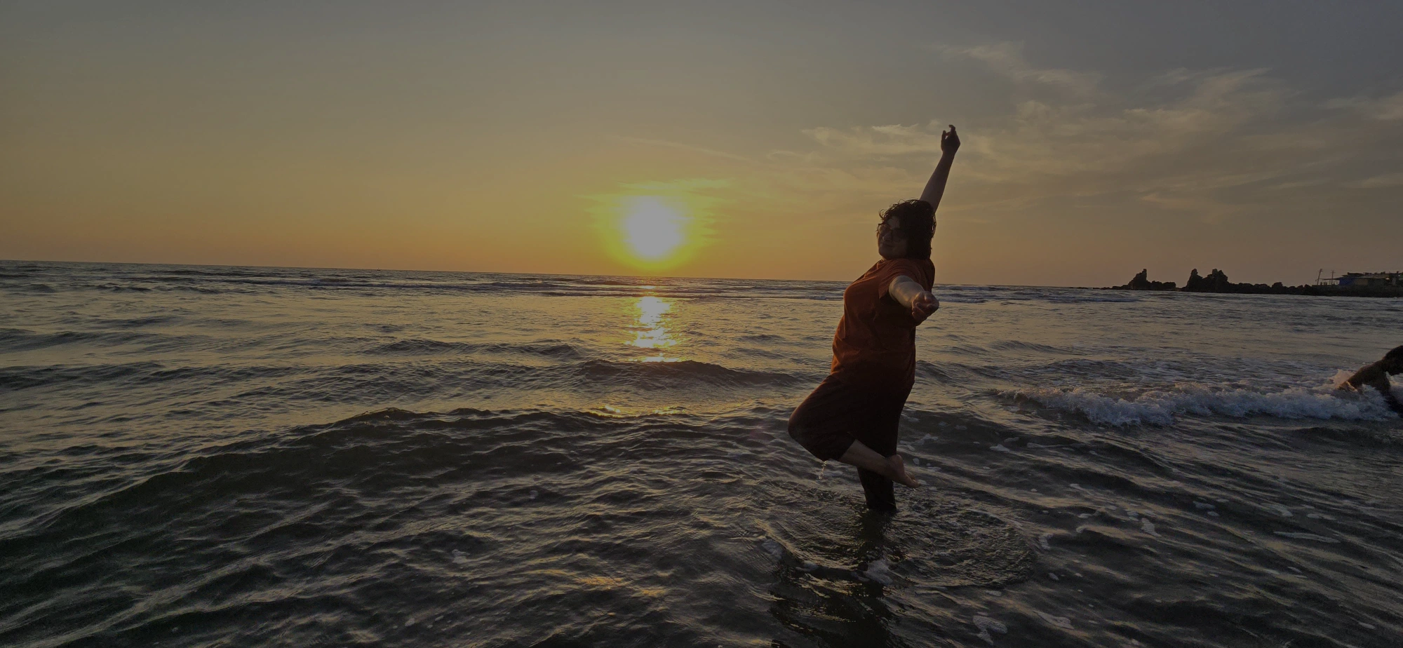 A person poses in a dance-like leap in shallow ocean water at sunset, one arm lifted toward the sky as golden light reflects across the waves.