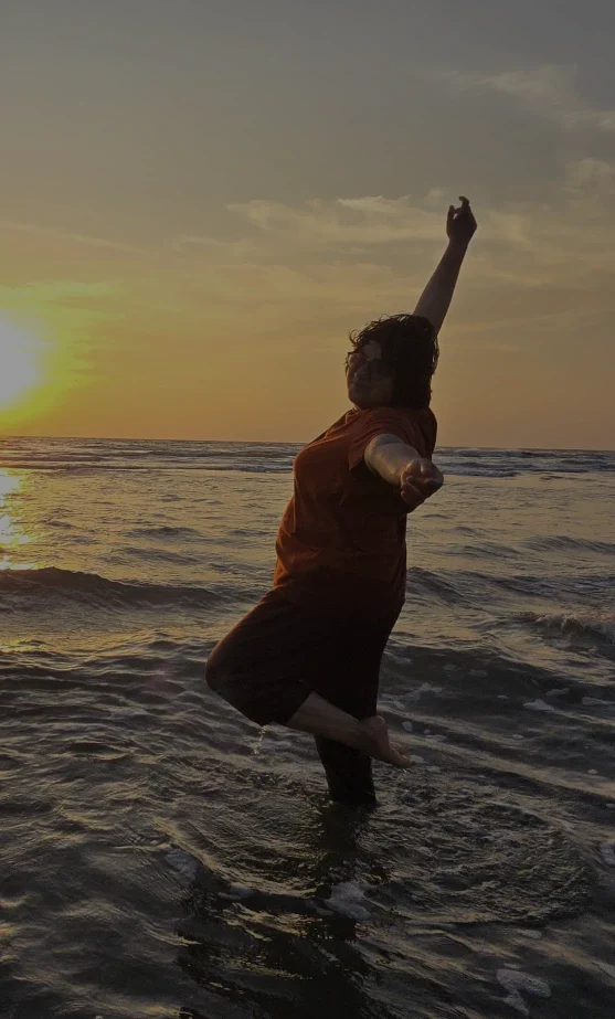 A person poses in a dance-like leap in shallow ocean water at sunset, one arm lifted toward the sky as golden light reflects across the waves.