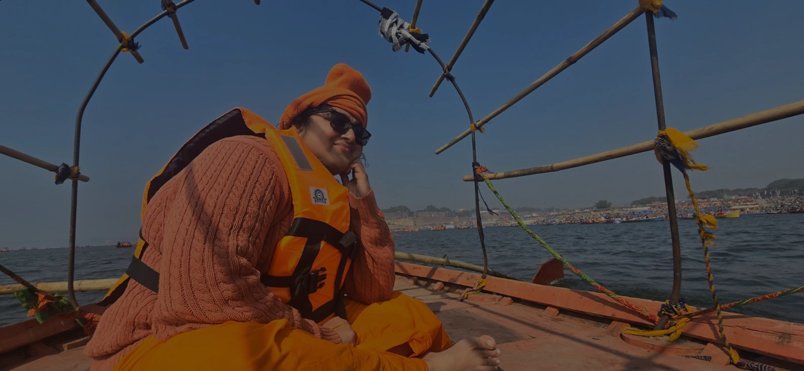 A person dressed in orange robes, an orange hat, and a bright safety vest sits cross-legged on a wooden boat, smiling. The boat is framed with bamboo arches, and a bustling riverside scene is visible across the water under a clear blue sky.