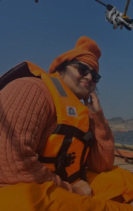 A person dressed in orange robes, an orange hat, and a bright safety vest sits cross-legged on a wooden boat, smiling. The boat is framed with bamboo arches, and a bustling riverside scene is visible across the water under a clear blue sky.
