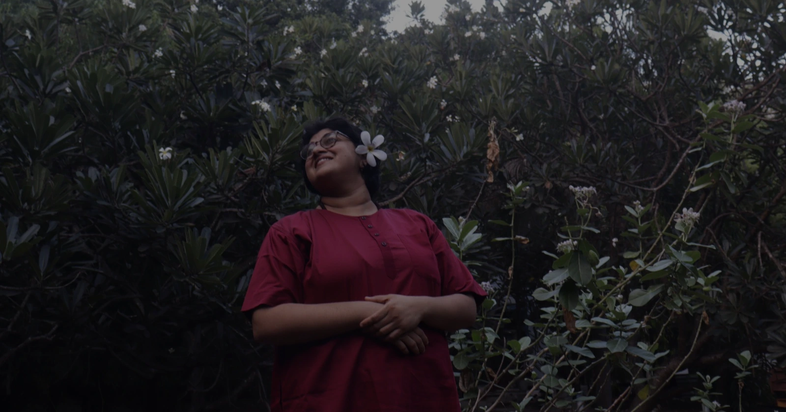 A person wearing a deep red outfit stands before dense green foliage, smiling softly with a white plumeria flower tucked behind their ear.