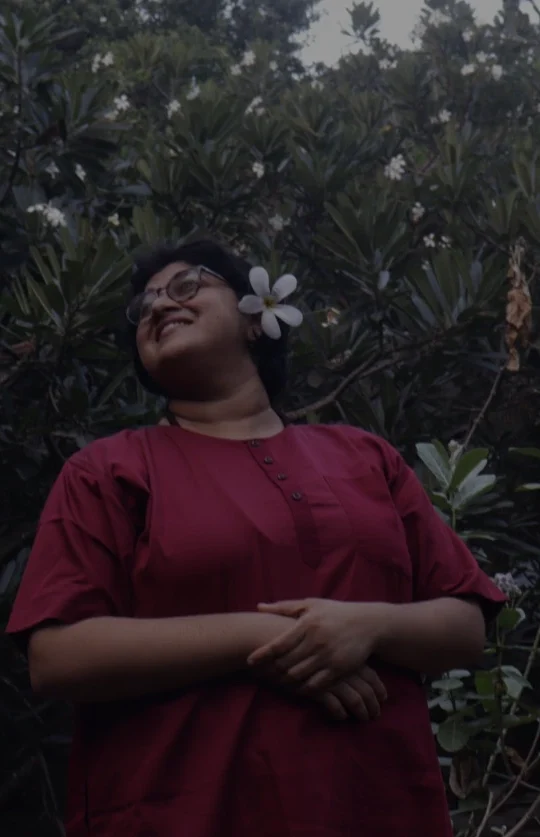 A person wearing a deep red outfit stands before dense green foliage, smiling softly with a white plumeria flower tucked behind their ear.