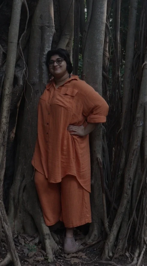 A person in a loose orange outfit stands among the massive twisting roots and aerial branches of a banyan tree in a shaded forest area, one hand on their hip.