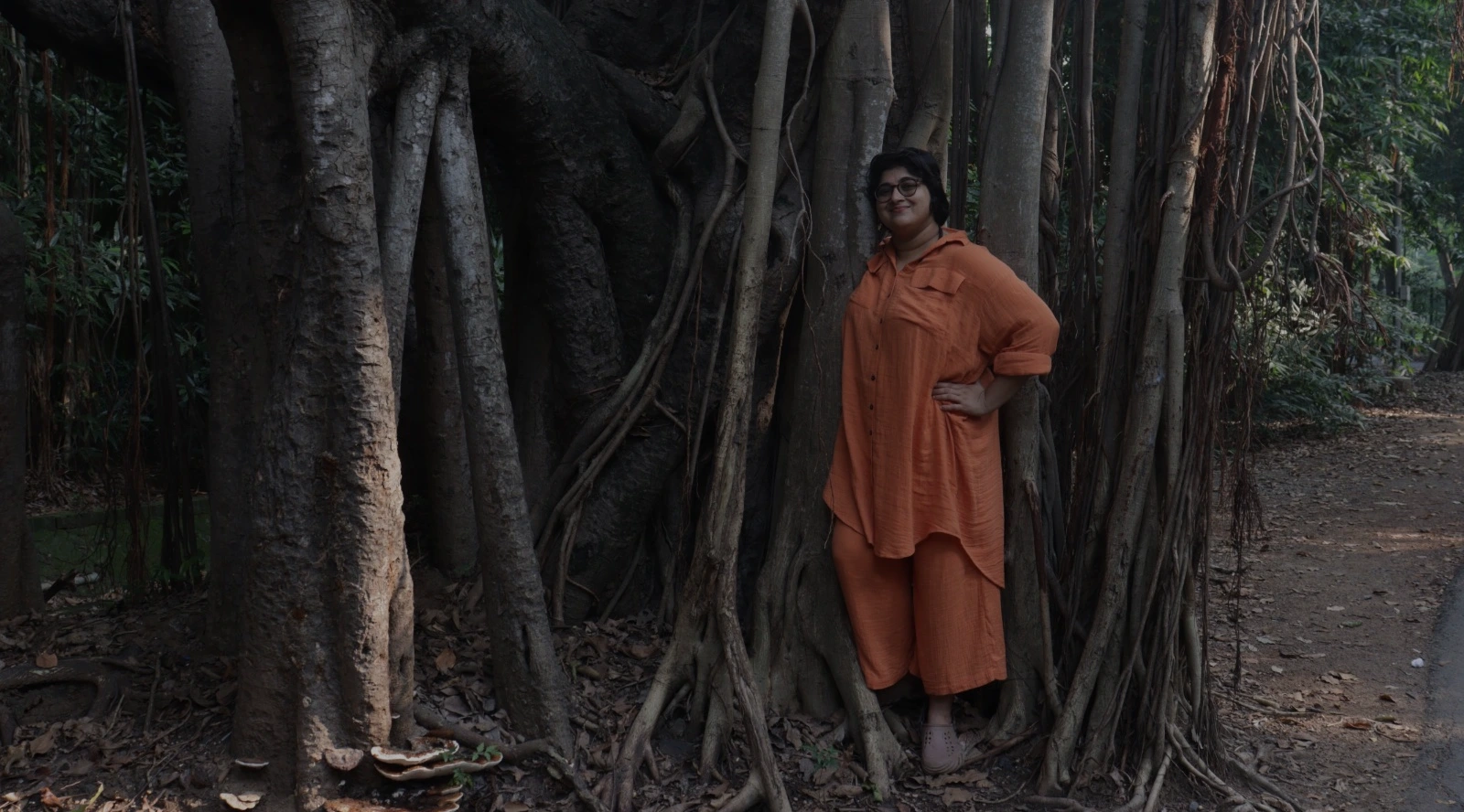 A person in a loose orange outfit stands among the massive twisting roots and aerial branches of a banyan tree in a shaded forest area, one hand on their hip.