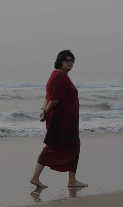 A woman walks barefoot along the shoreline, wearing a deep red outfit, with gentle waves rolling in behind her under a pale morning sky.