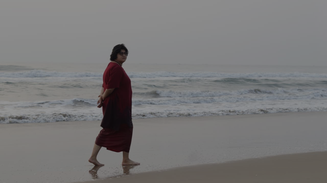 A woman walks barefoot along the shoreline, wearing a deep red outfit, with gentle waves rolling in behind her under a pale morning sky.