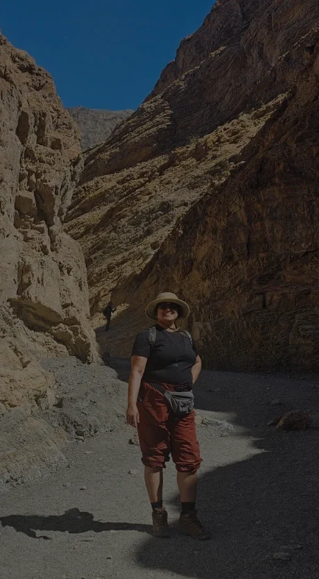 A person stands on a gravel path between two towering golden-brown canyon walls, wearing a hat, black shirt, and red pants under a bright blue sky.