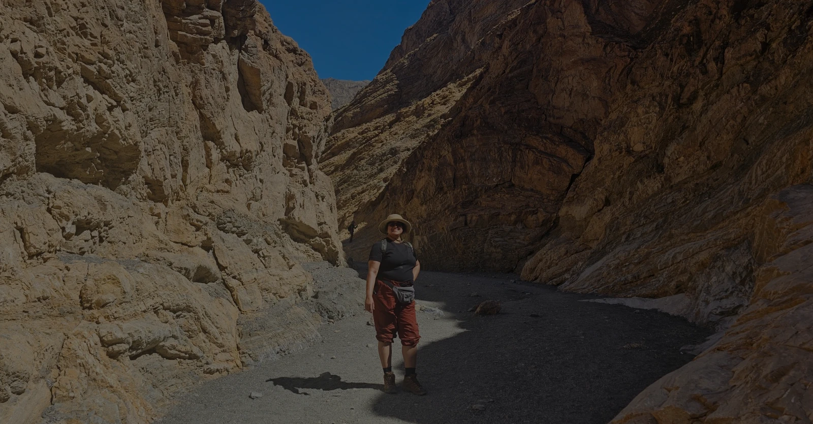 A person stands on a gravel path between two towering golden-brown canyon walls, wearing a hat, black shirt, and red pants under a bright blue sky.