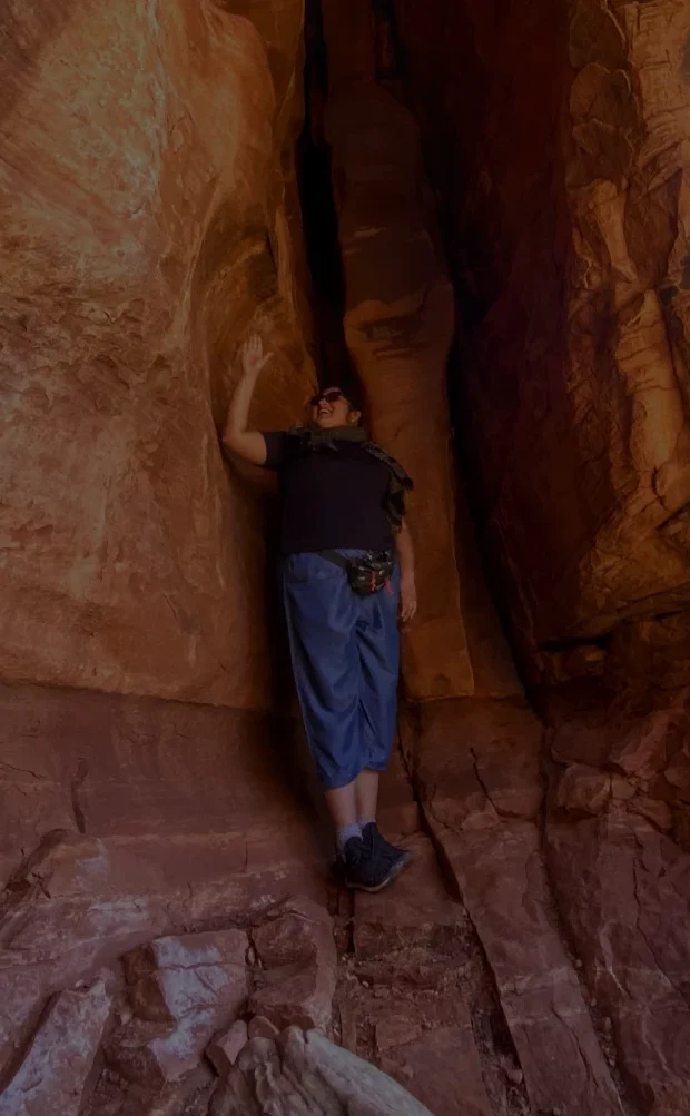 A person leans against the inside of a narrow red-rock crevice, surrounded by steep sandstone walls, with one arm touching the rock for balance.