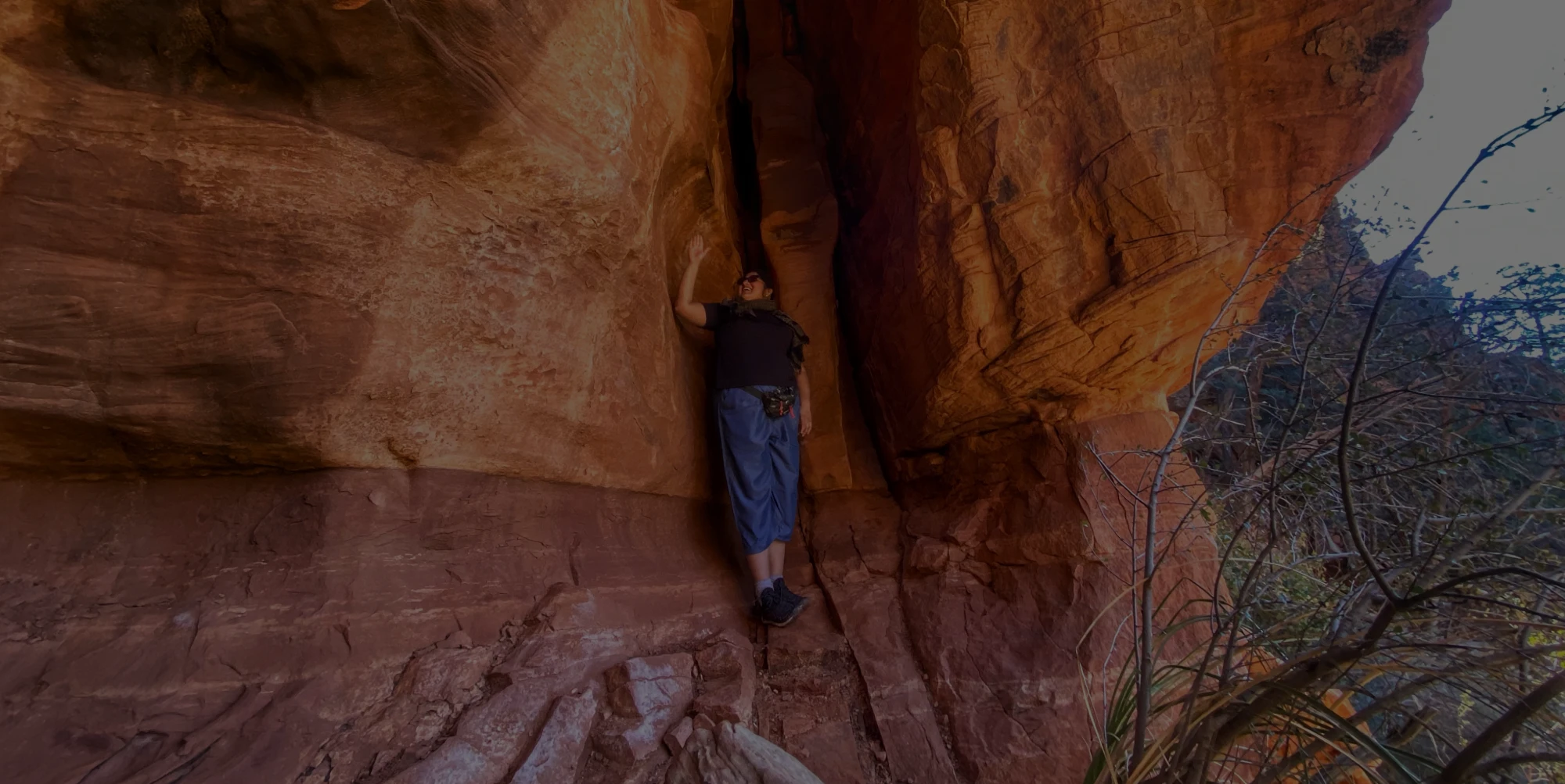 A person leans against the inside of a narrow red-rock crevice, surrounded by steep sandstone walls, with one arm touching the rock for balance.